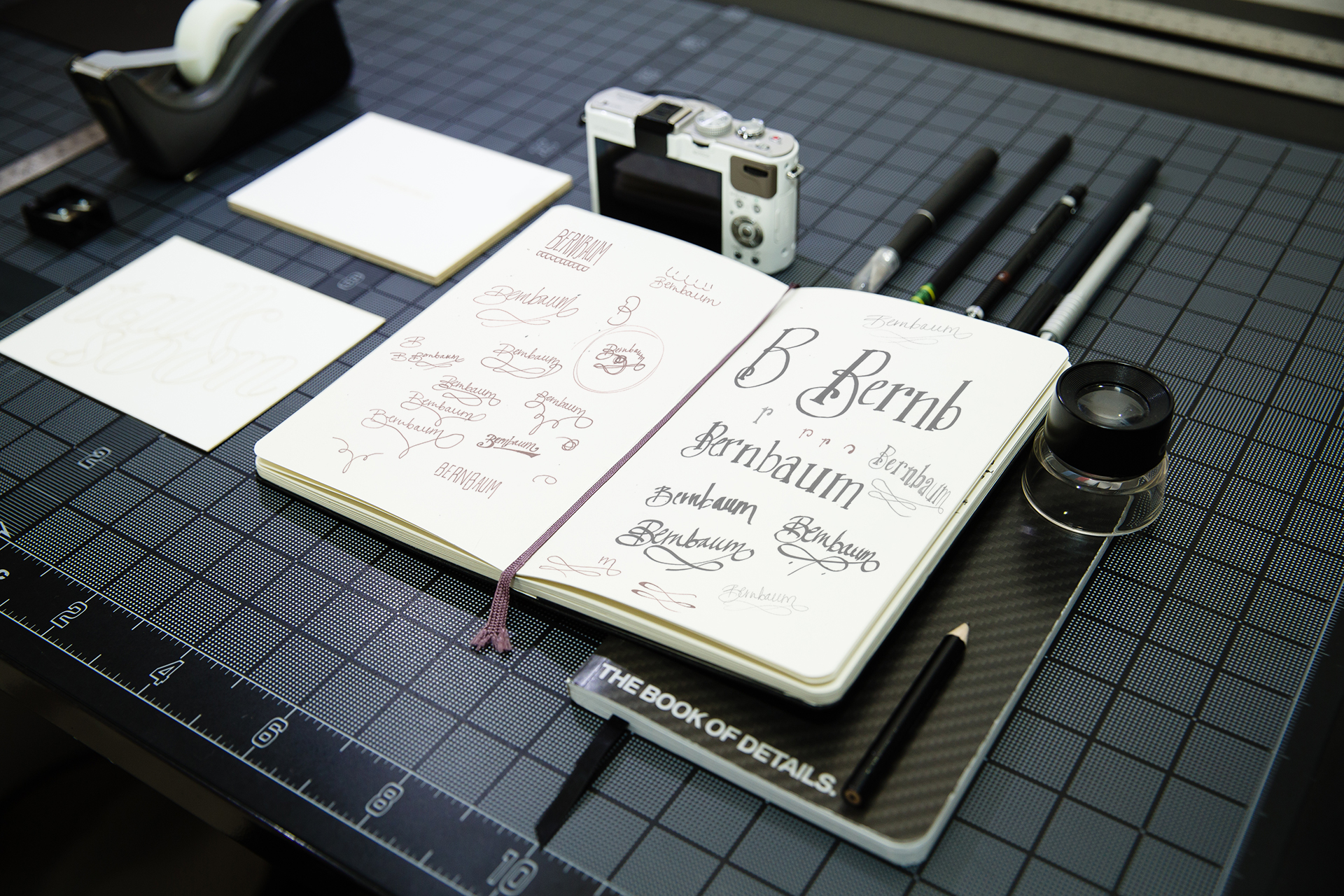 Sketchbook containing logo design concepts laying open on desk next to camera, and pens, and white card reading talk soon