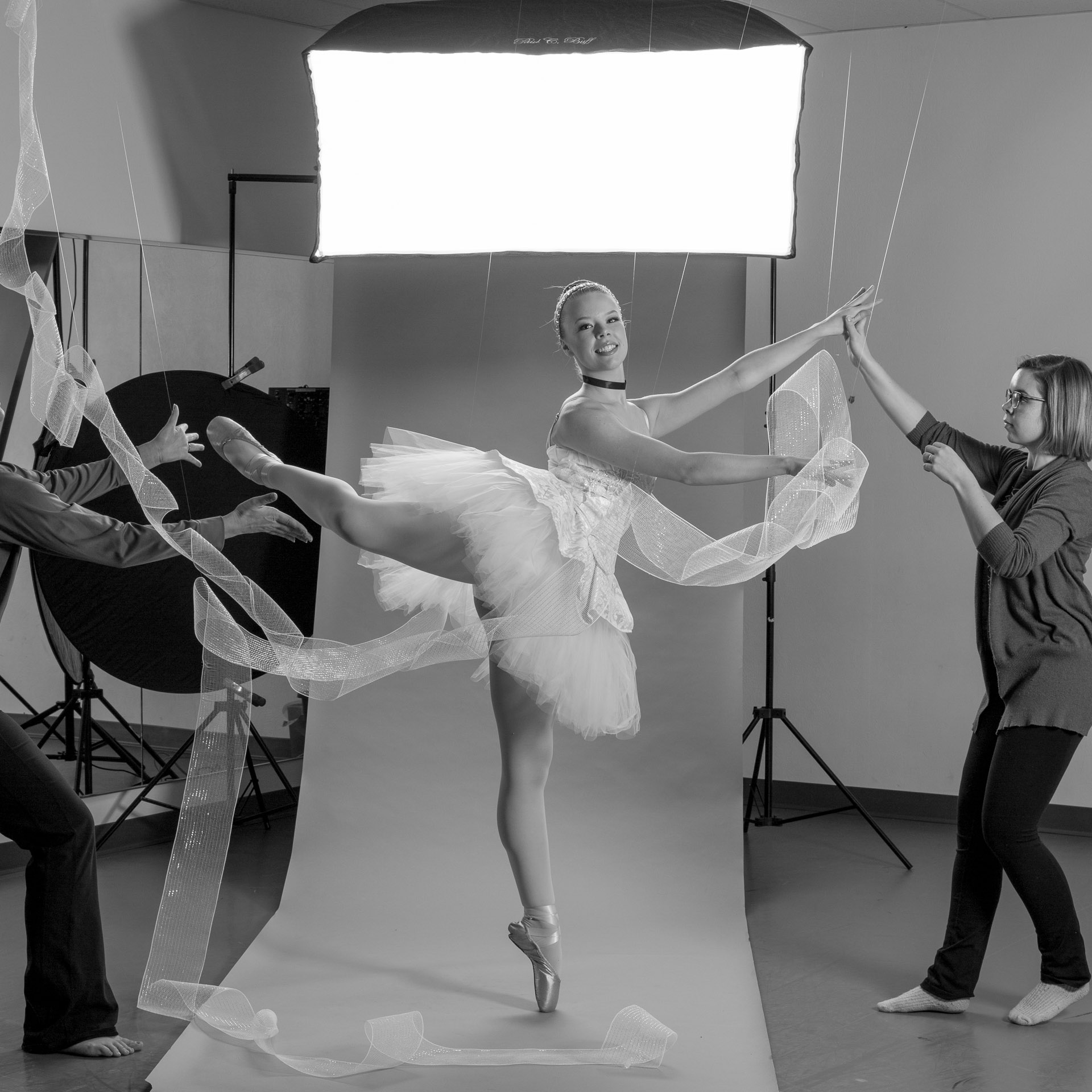 Ballerina dressed in Cinderella tutu poses in front of photo backdrop while two women hold her hand and foot