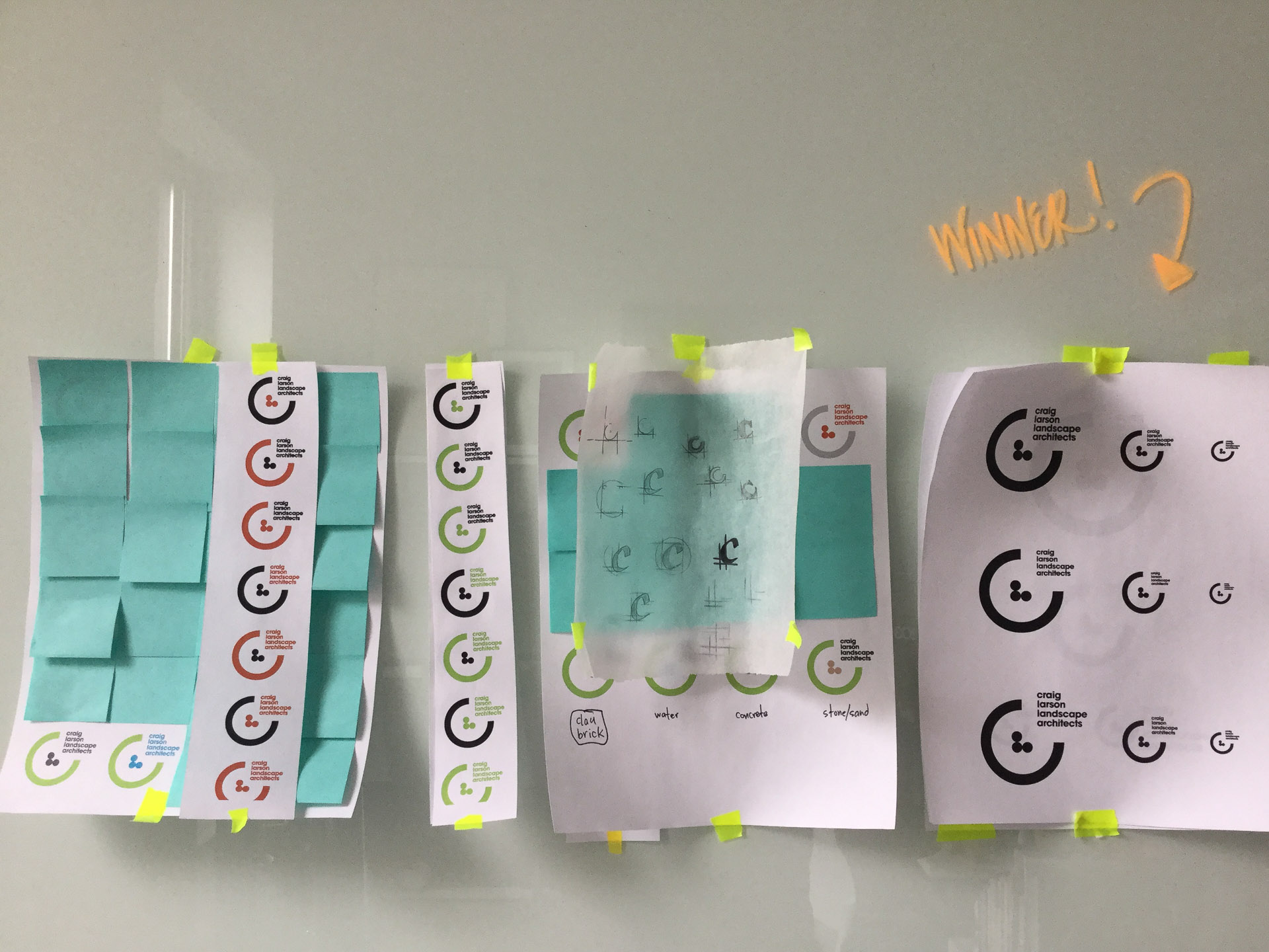 Logo concepts printed on letter paper and taped to a white board with blue post-its stuck on the papers