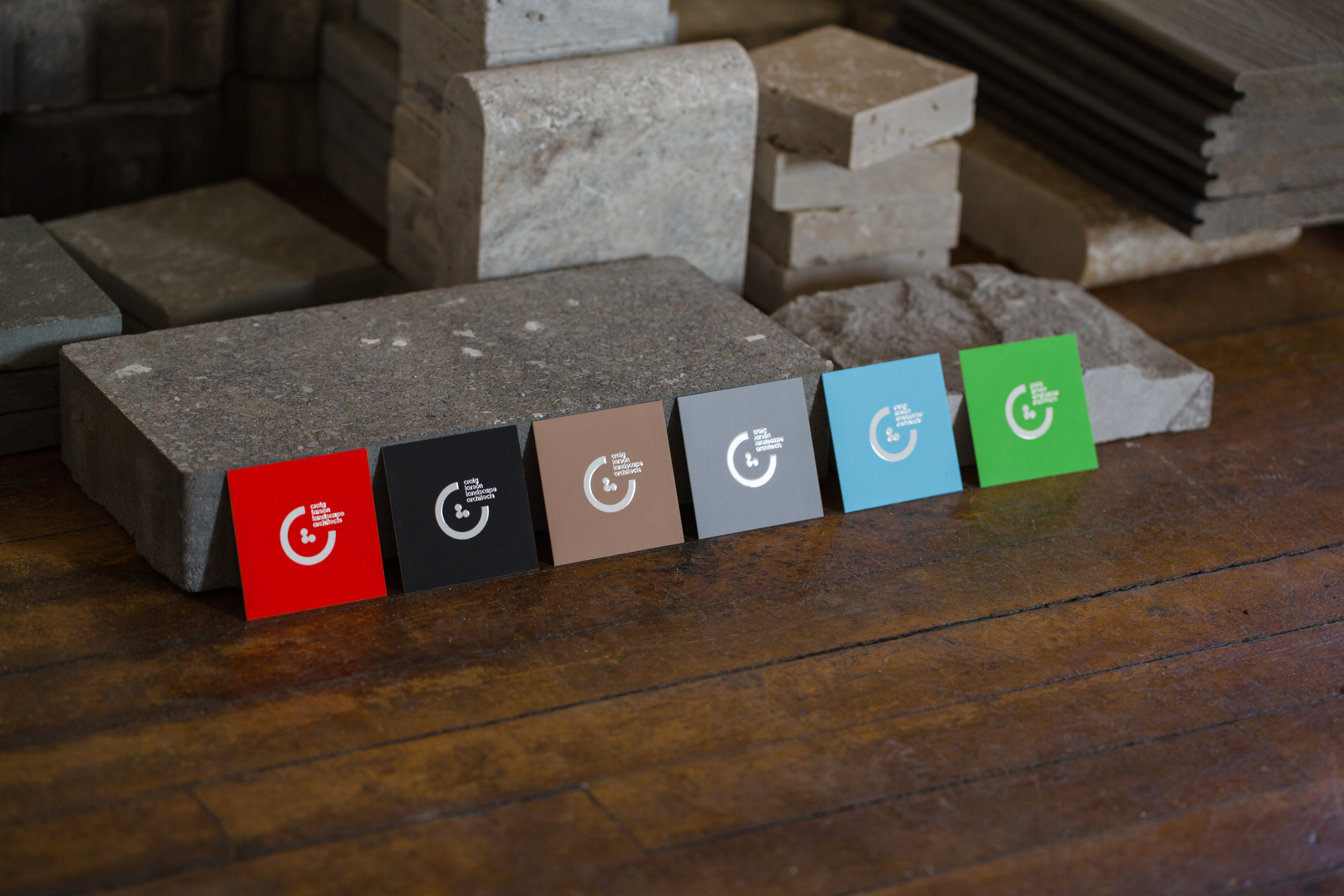 Red, black, tan, gray, blue, and green square business cards displaying CLLA logomark leaning up against stacks of stone