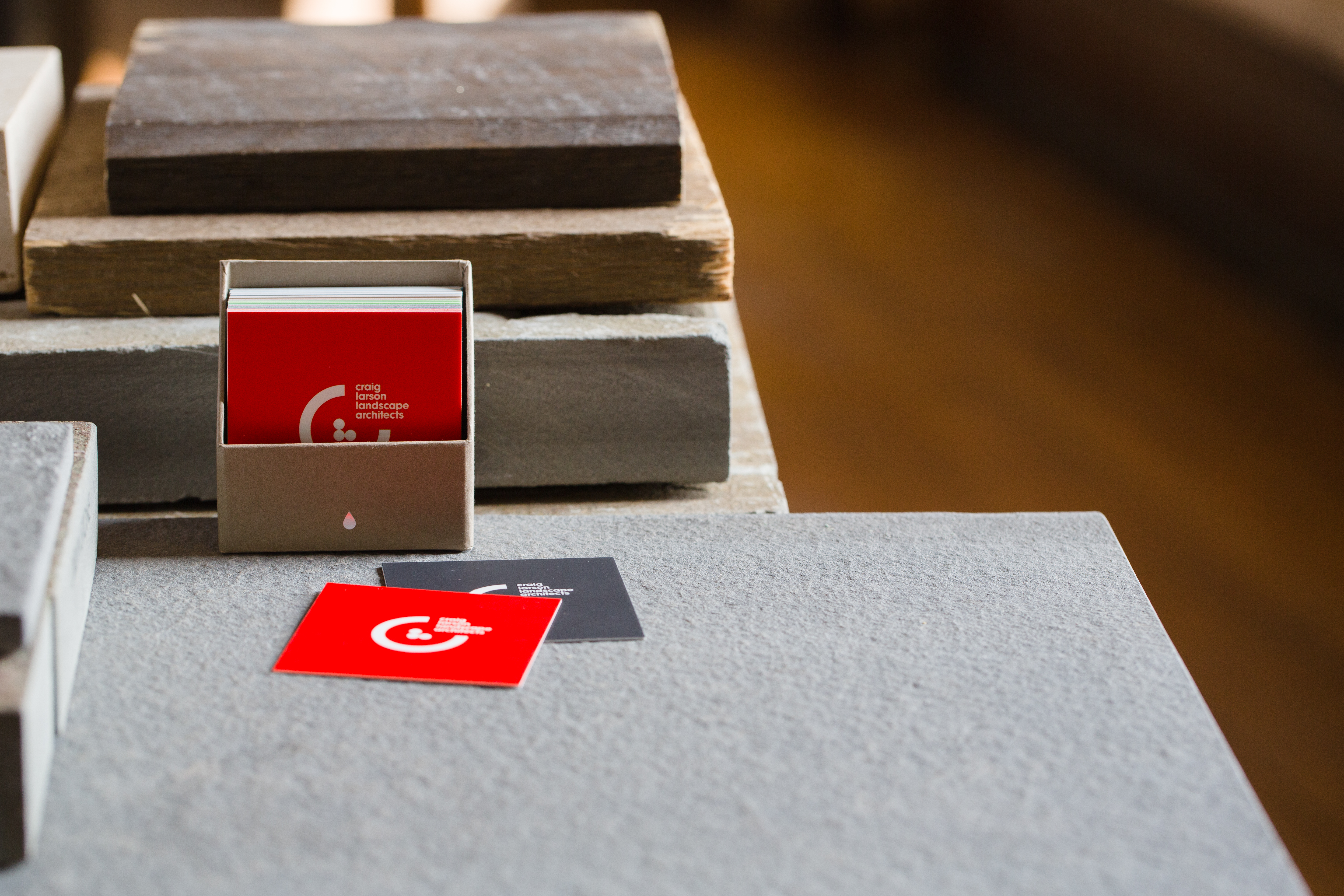 Box of red, branded, square business cards displaying the CLLA logomark sitting atop a stack of stone tiles