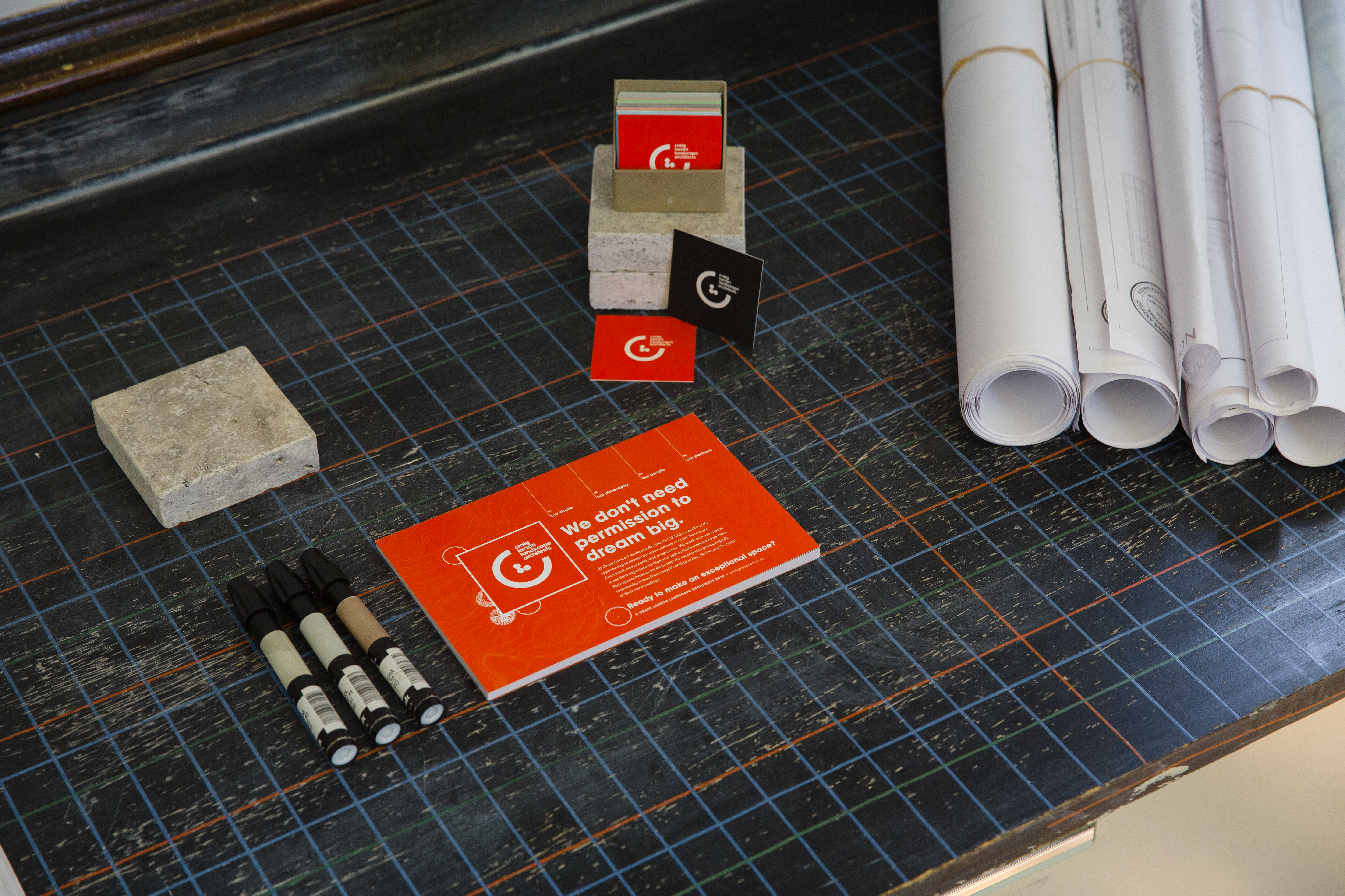 Red, CLLA-branded and box of square business cards next to rolled blueprints on a black gridded mat