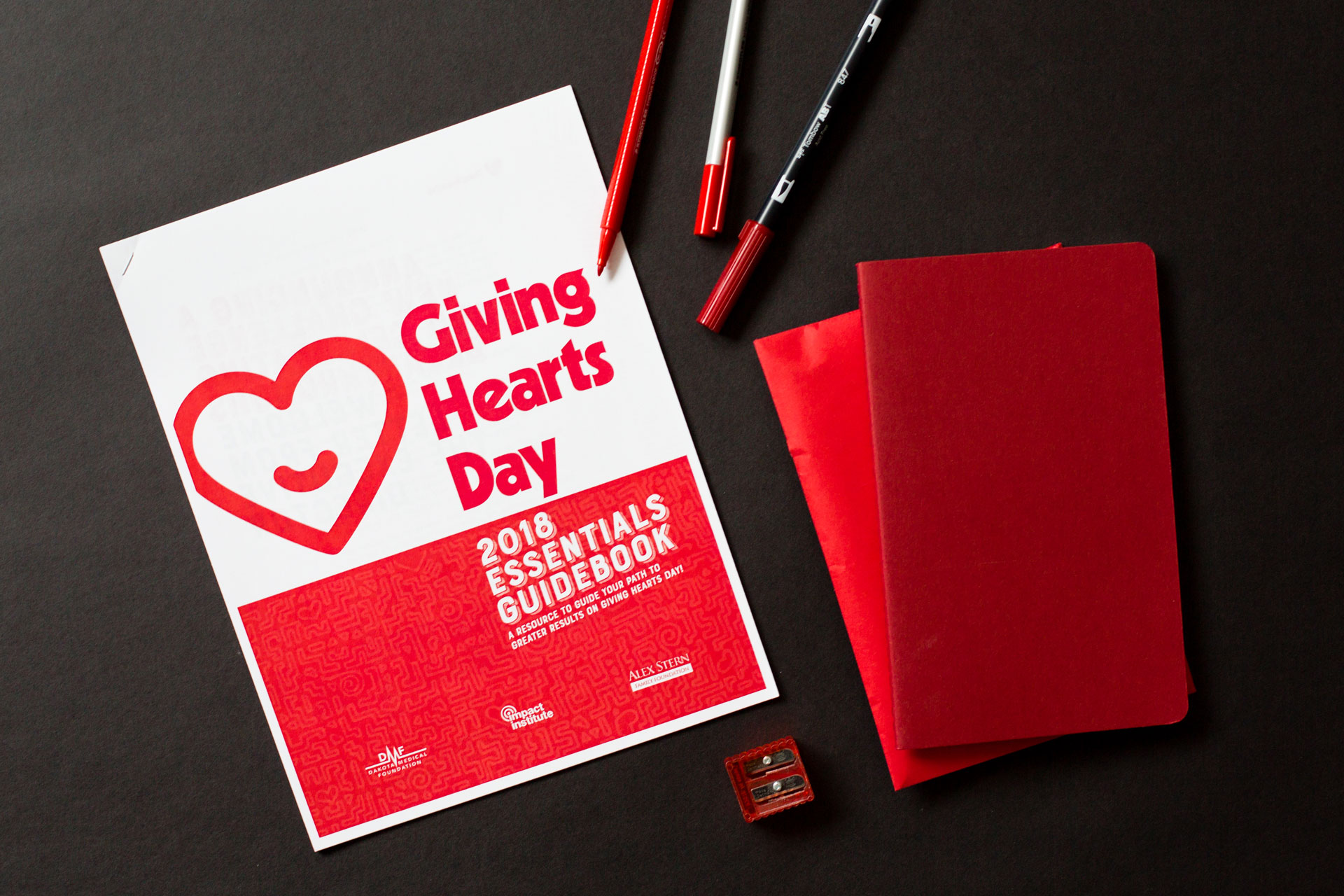 Giving Hearts Day 2018 Essentials Guidebook printed laying on a dark background next to red journals and markers