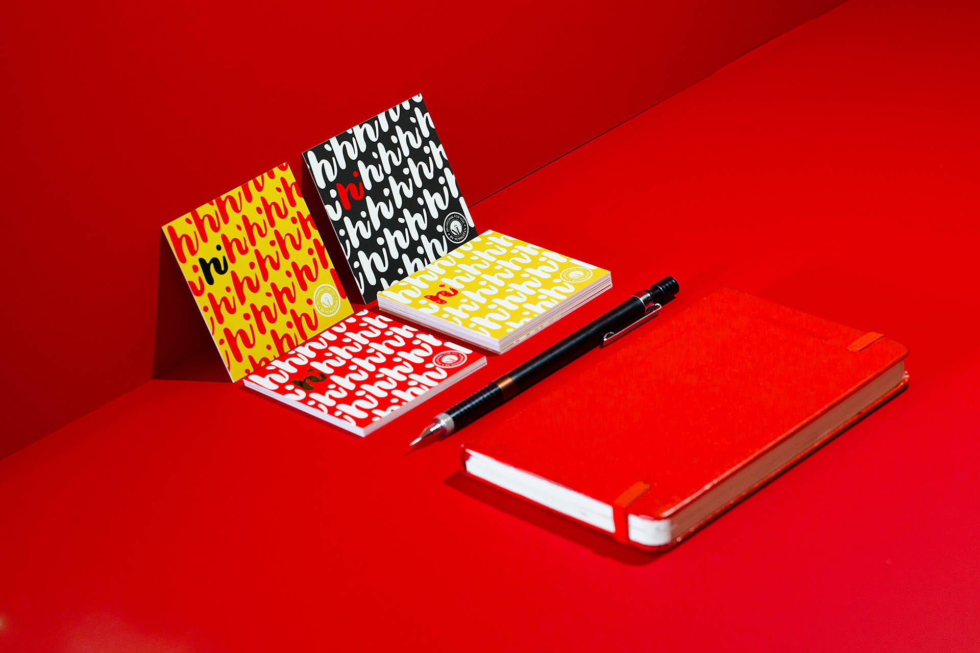 Four colorful cards covered with a repeating hi pattern, a red journal, and black pen on a red background