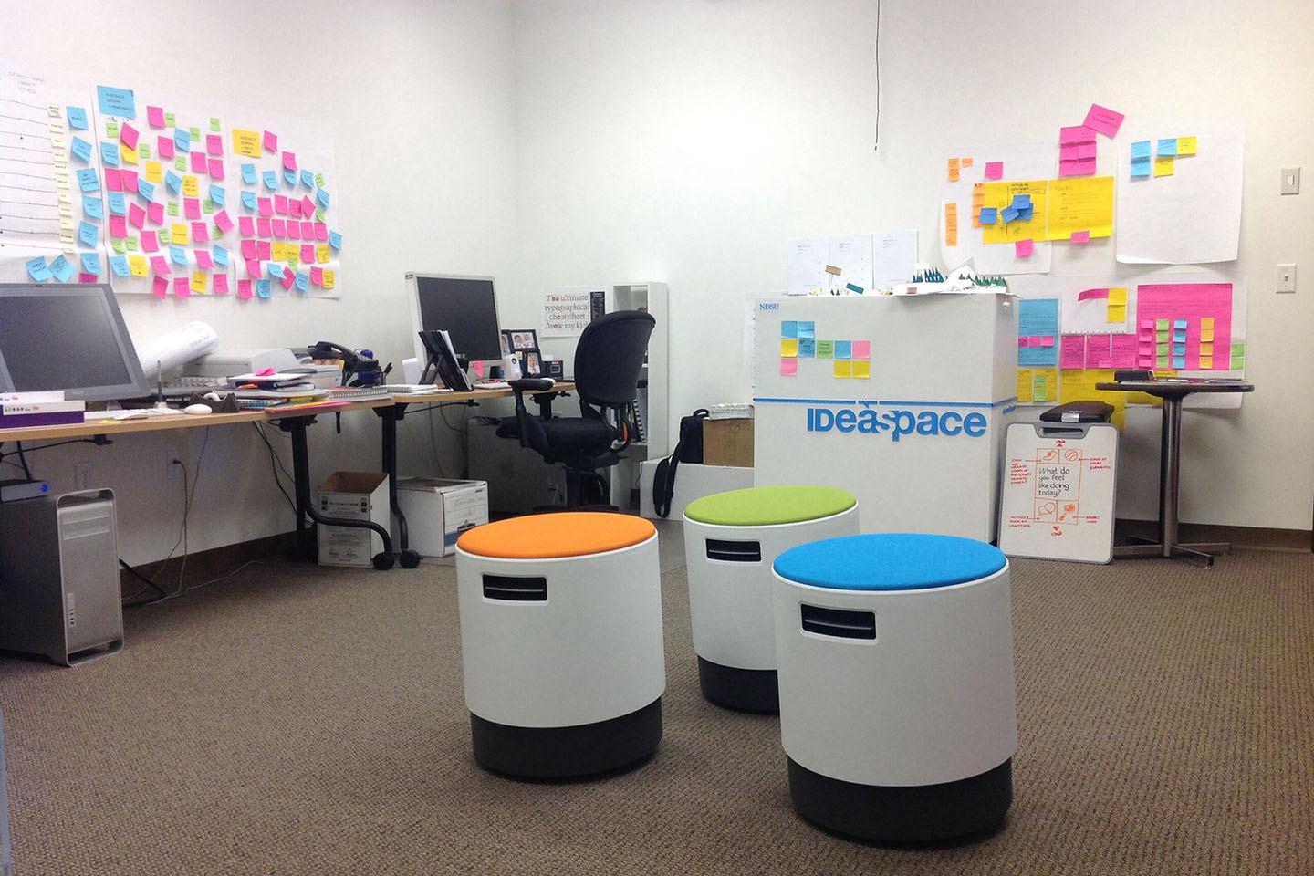 Colorful stools, post-it notes, and two desks in an office with white walls and brown carpet