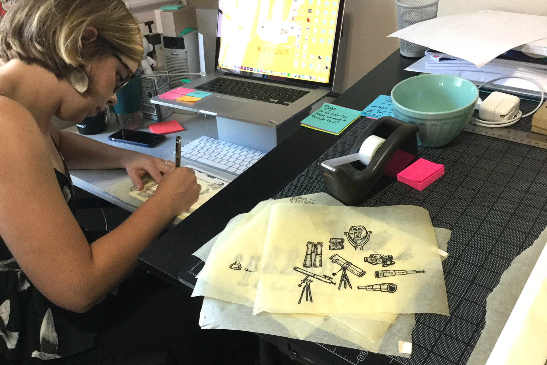 Woman drawing with pieces of tracing paper displaying illustrations of telescopes stacked beside her on a desk