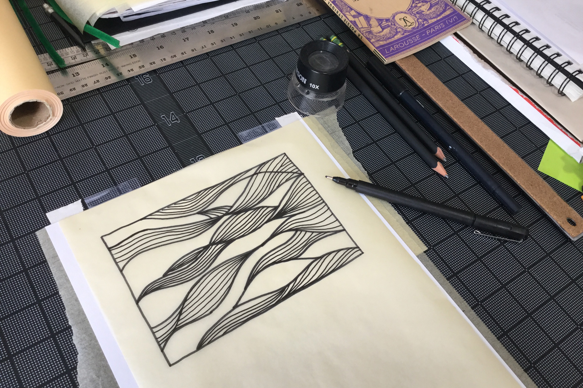 A wavy pattern hand-drawn on a sheet of tracing paper next to pens and notebooks atop a black gridded mat