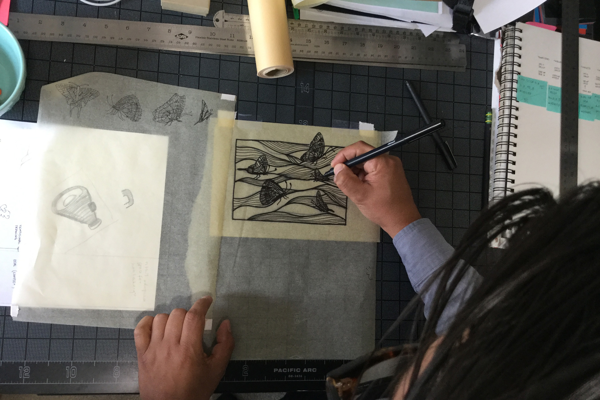 A man drawing waves and butterflies on a piece of tracing paper atop a black gridded mat