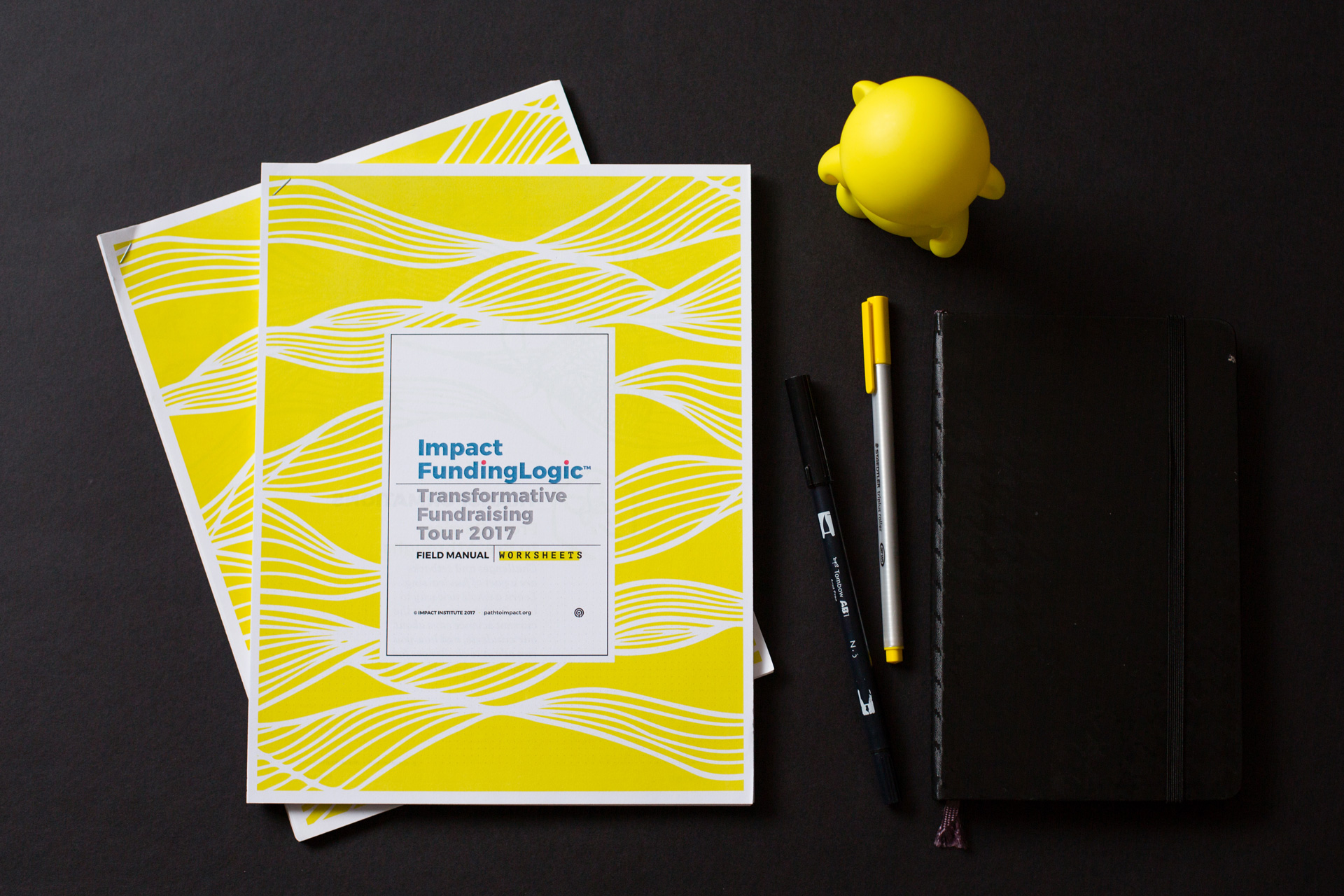 Printed Impact FundingLogic packet with yellow cover next to pens and a yellow munny figurine on a black background