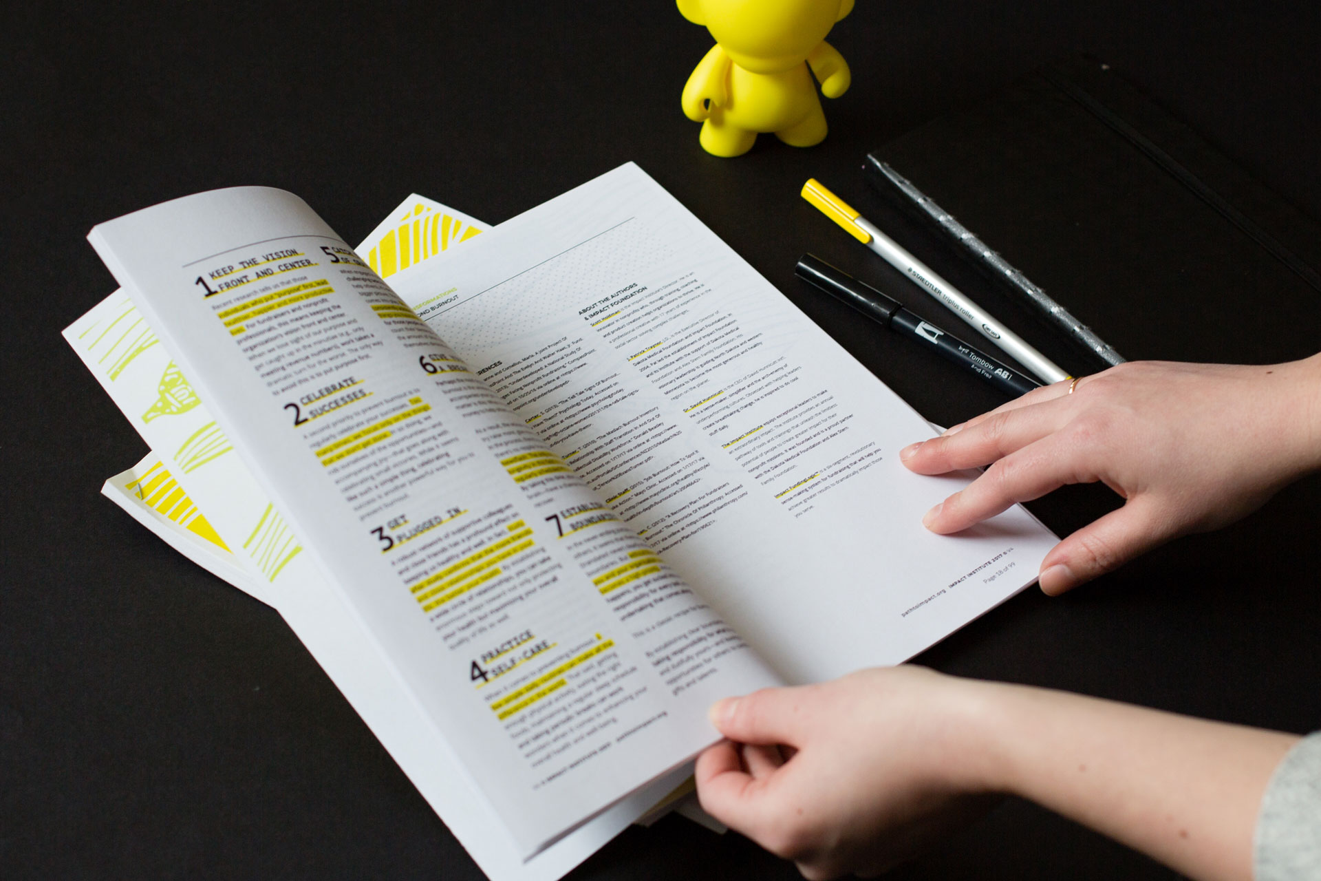 Two hands holding printed materials open next to pens and a yellow munny figurine on a black background