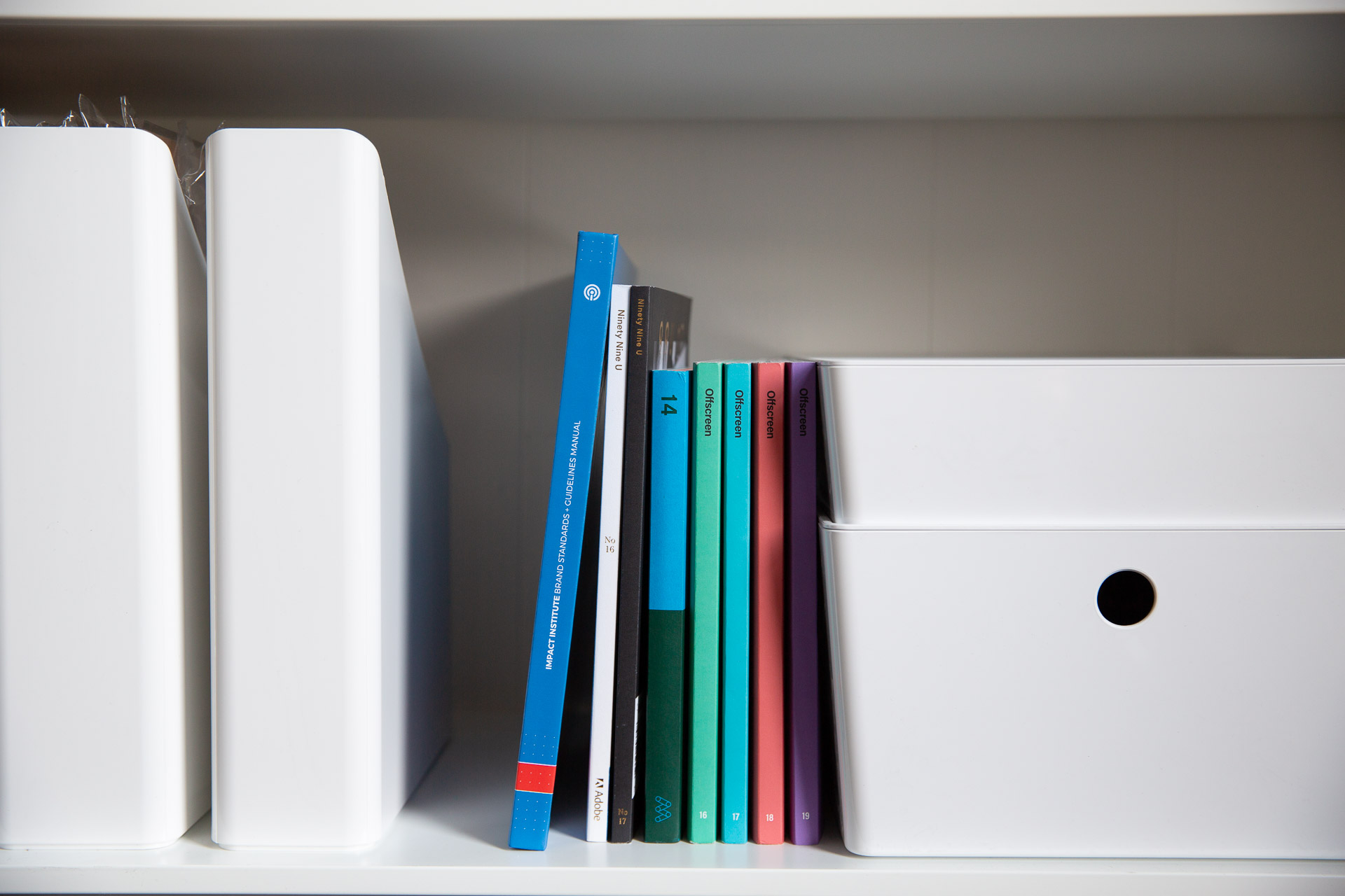 Impact Institute brand guideline manual stacked on bookcase next to small, colorful magazines and white bins