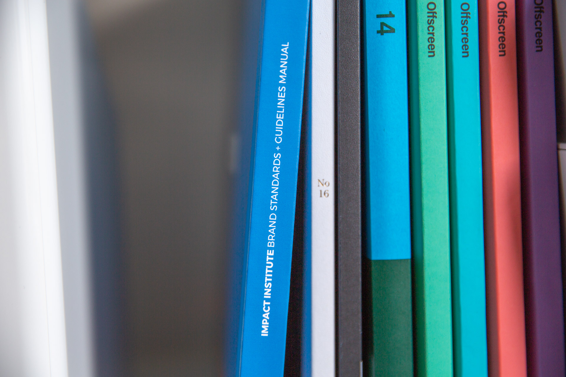 Close-up of Impact Institute brand guideline manual stacked on bookcase next to small, colorful magazines and white bins