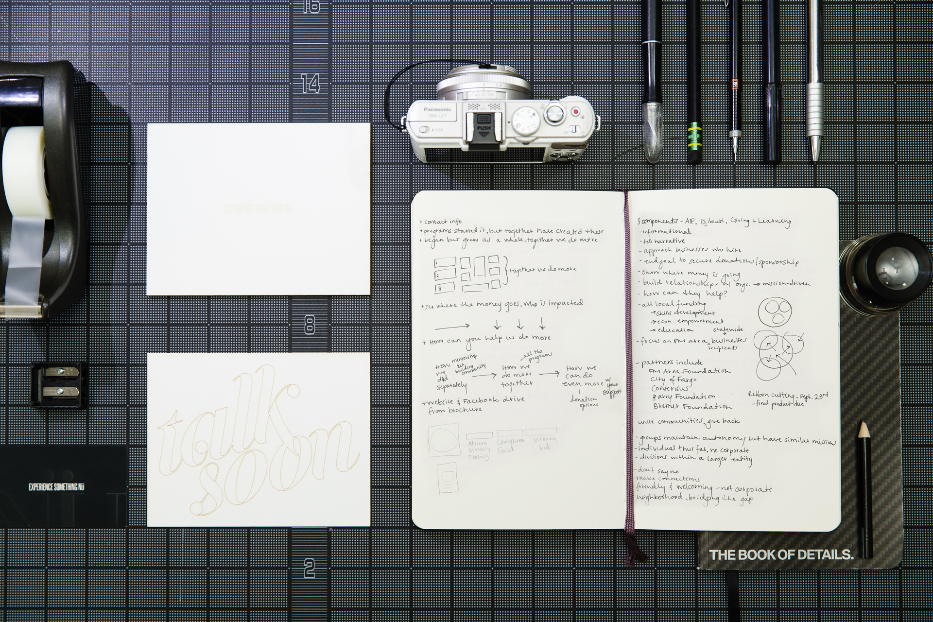 Open sketchbook full of sketches next to cards, tape dispenser, and pens on black gridded background