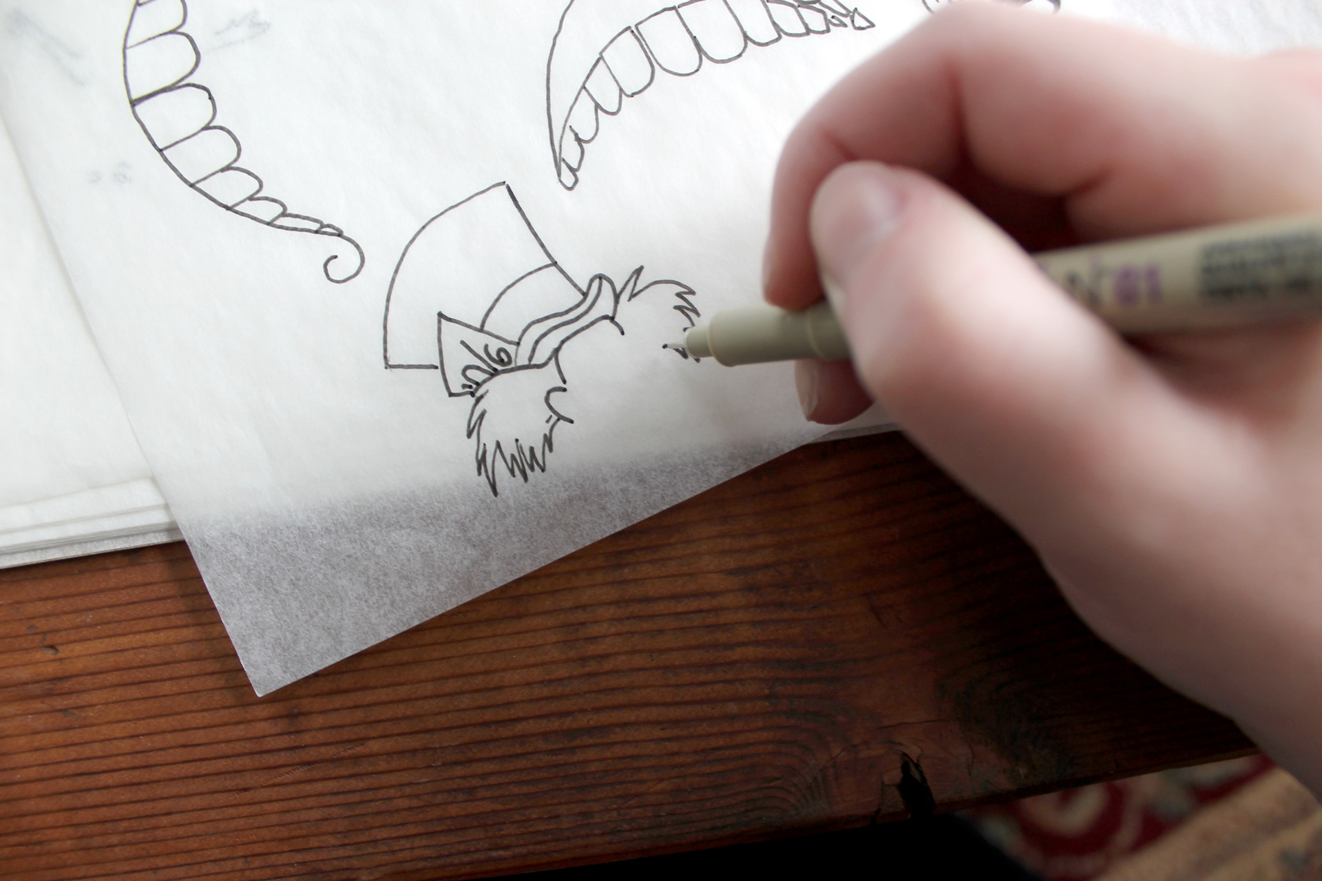 Close-up of hand drawing Mad Hatter illustration on tracing paper atop a wooden table