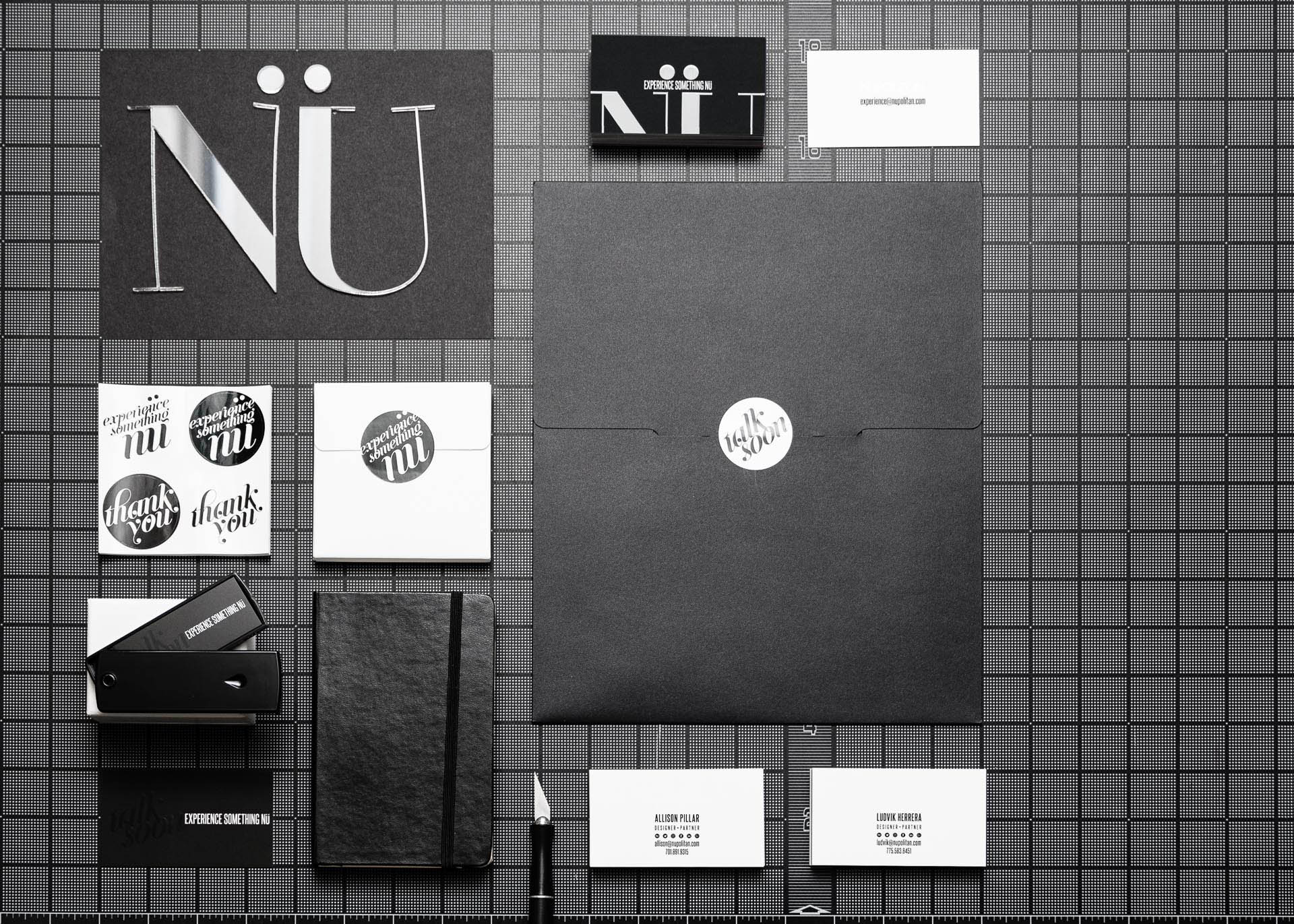 NÜ in acrylic letters, black and white round stickers, business cards, and a large black envelope on a black gridded surface