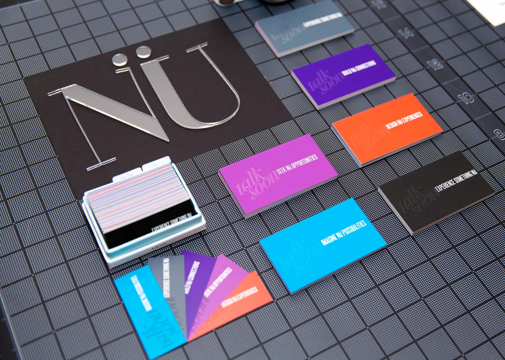 NÜ in acrylic letters, a white camera, and purple, blue, and red business cards on a black gridded surface