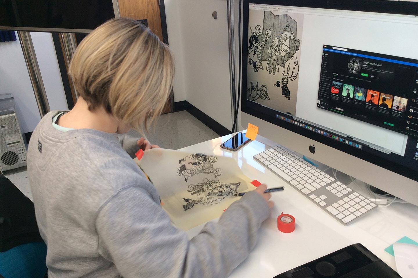 Woman in gray sweatshirt drawing on tracing paper atop a white desk