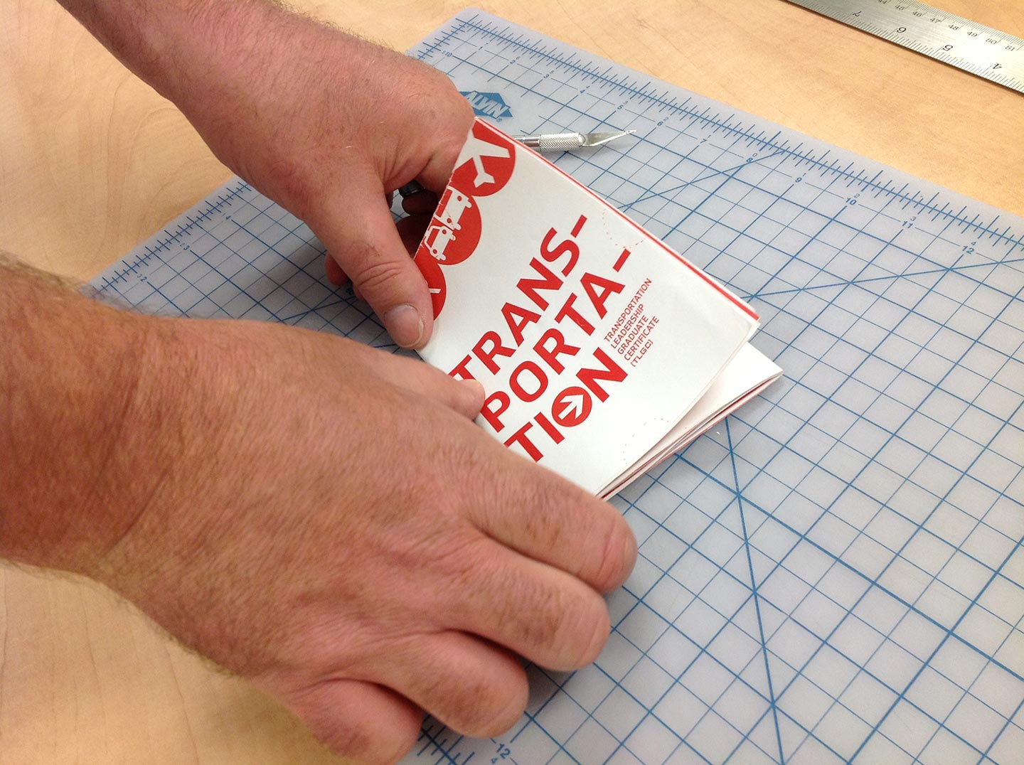 Two hands folding a transportation brochure atop a light blue gridded cutting mat