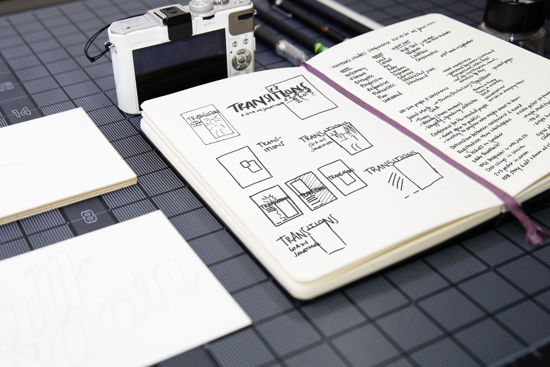 Sketchbook of design concepts next to a white camera and white cards atop a black gridded surface