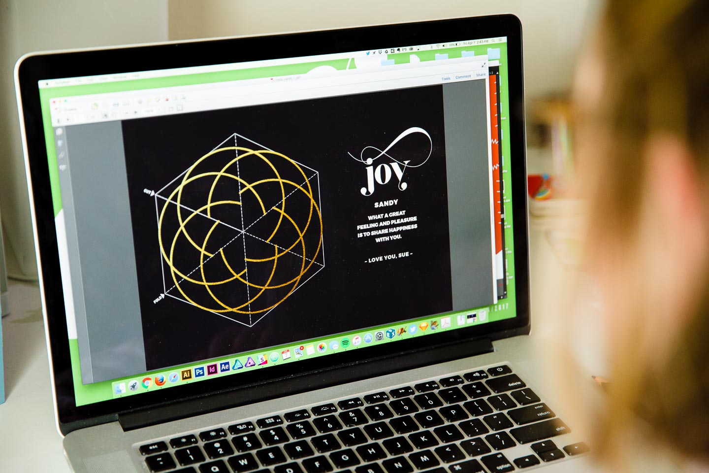 A woman working on a laptop creating a black postcard that reads Joy next to a circular pattern